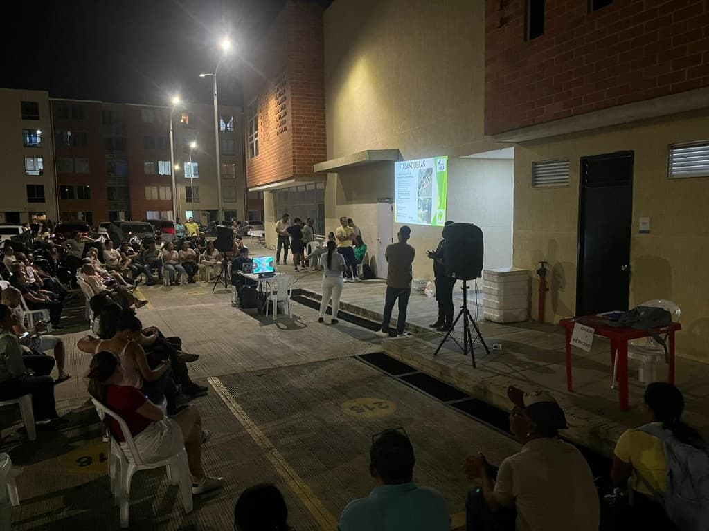 Reunión comunitaria con asistentes en sillas, equipo de sonido y conjunto residencial al fondo